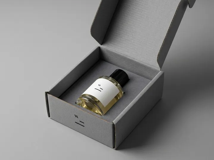 Clear cylindrical perfume bottle seated in a fitted insert inside an open gray mailer box