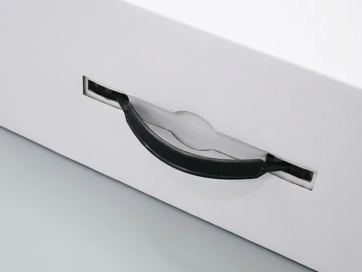 Close-up of a black plastic carry handle mounted through two slots in a white corrugated hat box panel