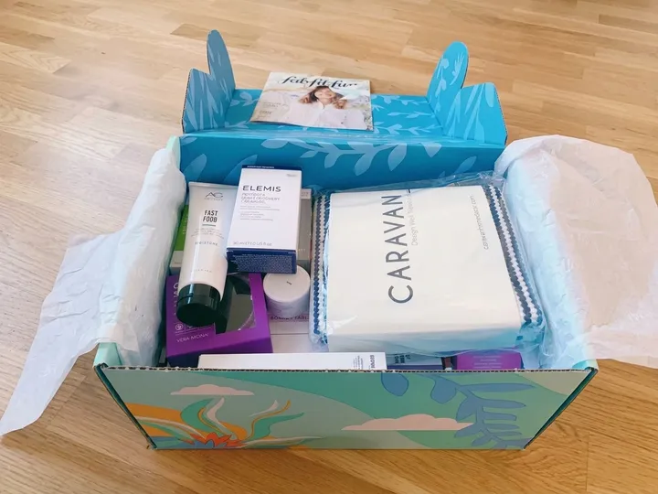 Open printed monthly subscription box holding assorted skincare, wellness, textile, and card items with tissue