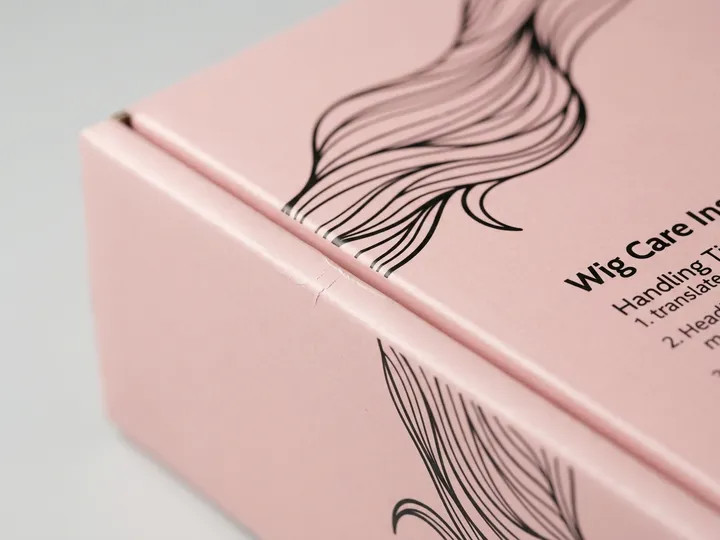 Close view of black hair artwork, pink offset print, gloss surface, score, and folded edge on a wig mailer box