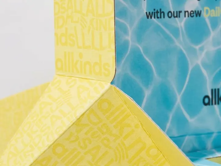 Close view of yellow repeat-letter print and blue water-pattern print meeting across folded corrugated mailer panels